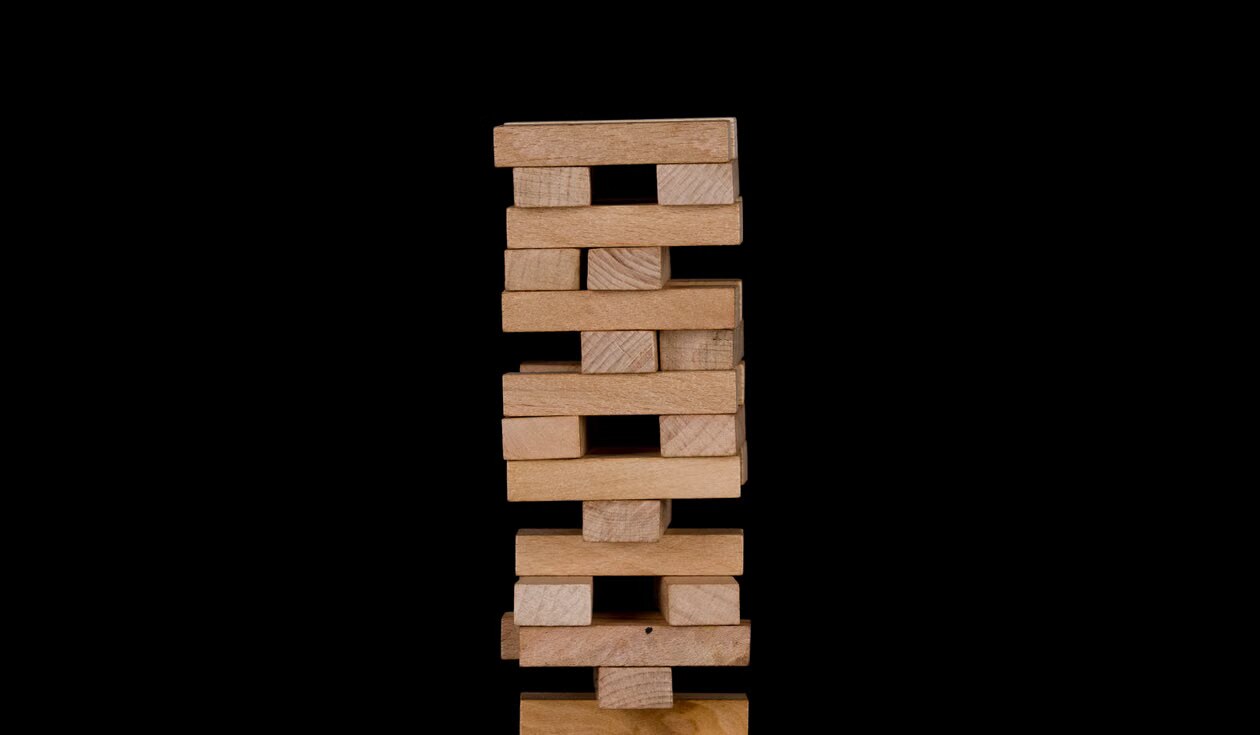A tower of wooden blocks stacked high against a black background, leaning and close to toppling